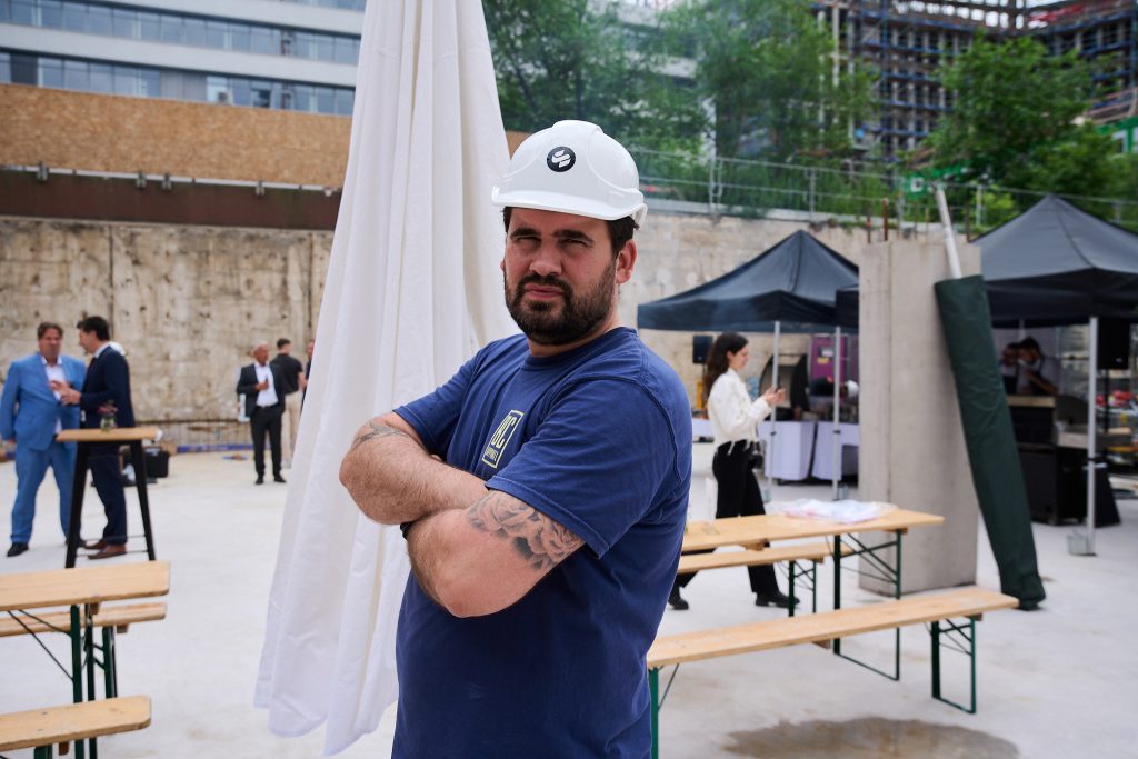 A construction worker wearing a white hard hat and a blue t-shirt stands with arms crossed, looking confidently at the camera. The setting appears to be a construction site with unfinished structures in the background. The worker's tattooed arms and determined expression highlight a sense of pride in his work. Behind him, people in business attire engage in conversation, creating a contrast between the formal and industrial elements of the scene. The environment suggests a moment during a construction project or event.