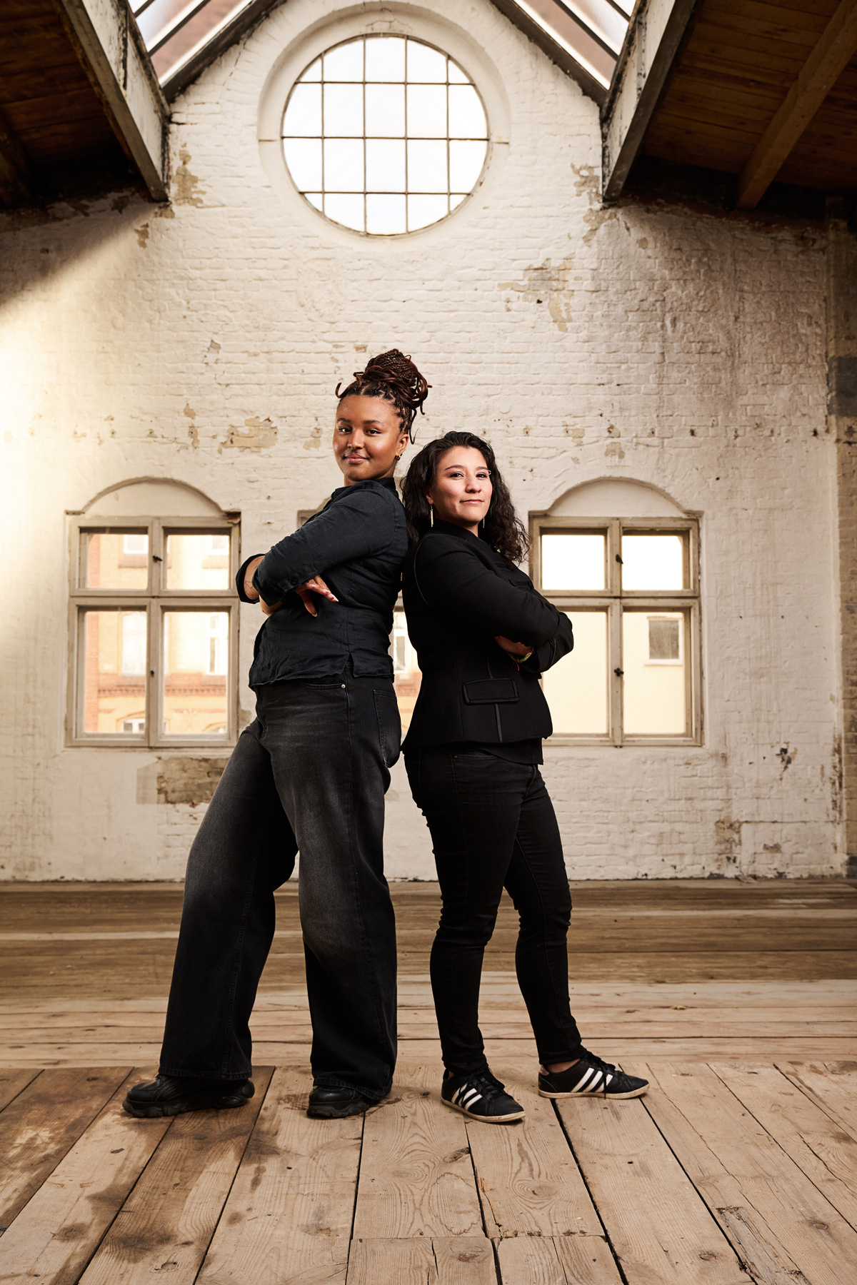 Two confident women stand back-to-back with arms crossed, in an industrial-style room with rustic wooden floors and weathered white brick walls. The space features large arched windows that allow natural light to pour in, highlighting the duo's strong, determined expressions. The women are dressed in dark, casual attire, with one sporting long braids and the other with wavy hair, reflecting a sense of empowerment and partnership.