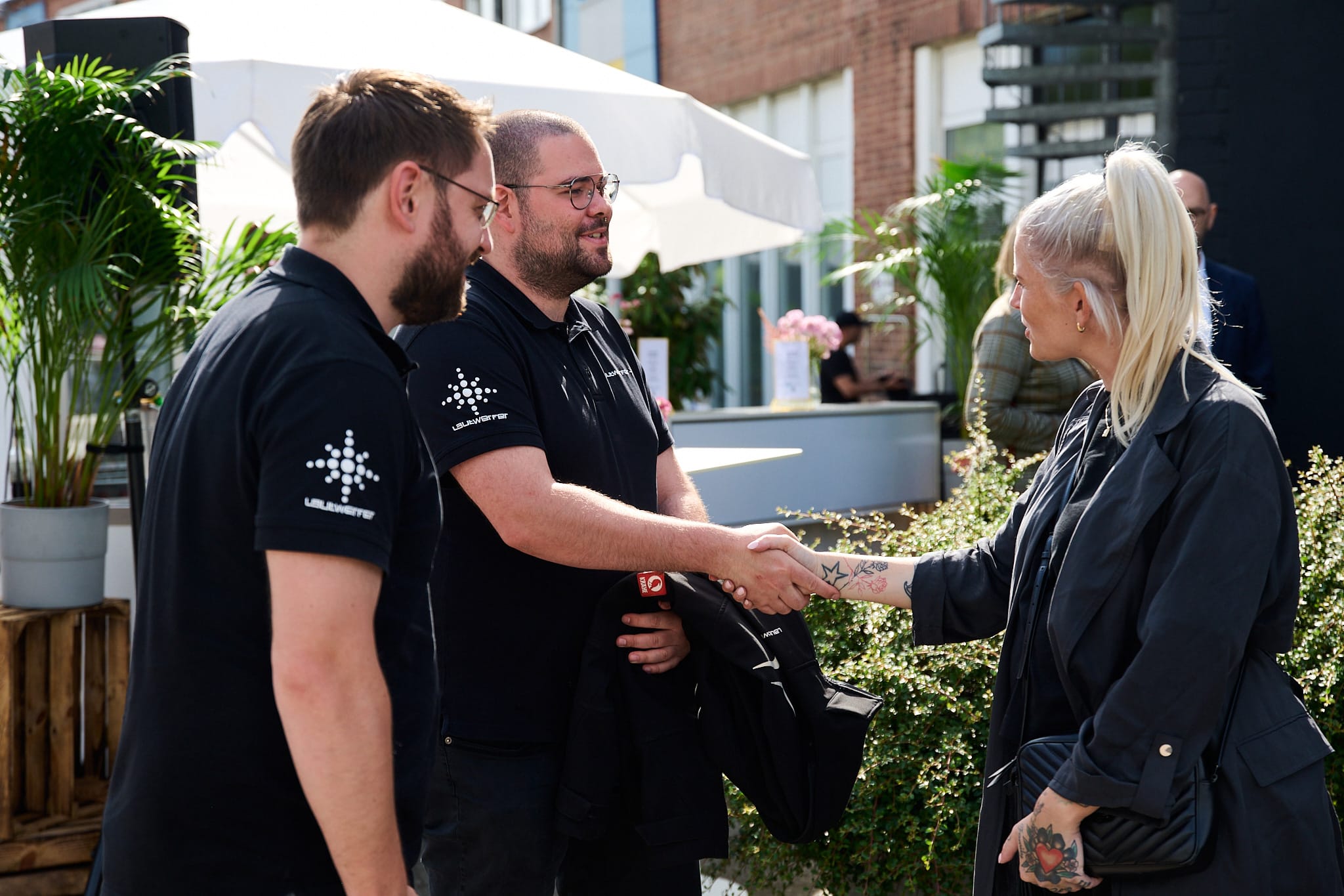 About Us 3 Drei Personen begrüßen sich bei einer Outdoor-Veranstaltung mit Händedruck und Lächeln.