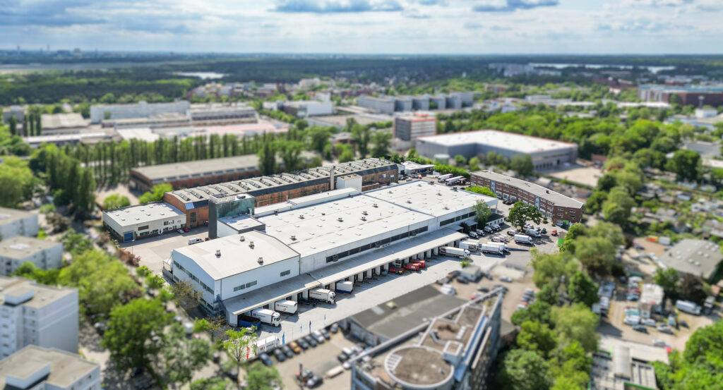 Leasing - Feevr 1 Luftaufnahme eines Logistikzentrums mit umliegenden Gebäuden und grünem Stadtgebiet.