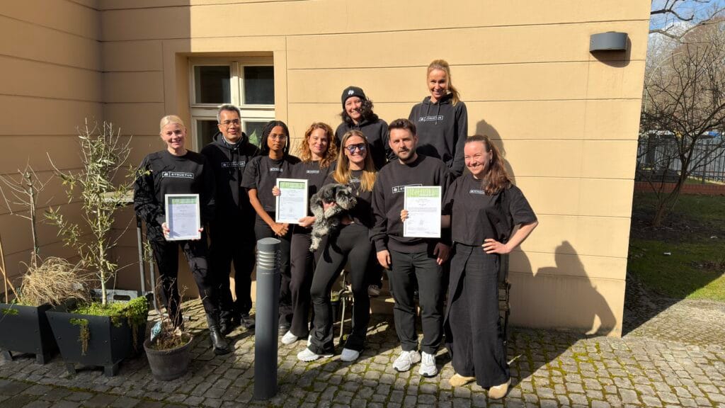 Teamfoto mit Zertifikaten und einem Hund, vor einem Gebäude im Freien.
