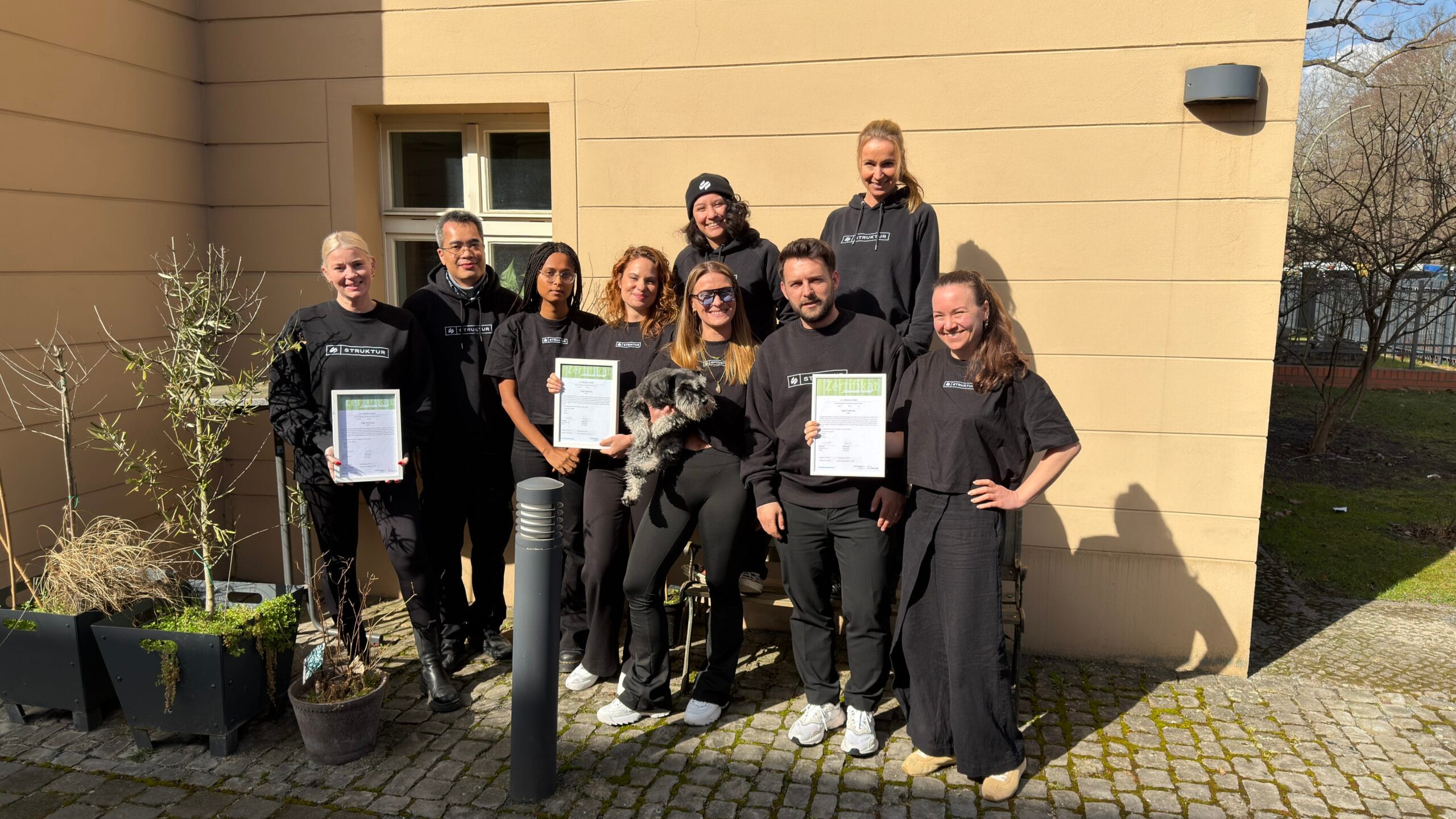 About Us 1 Teamfoto mit Zertifikaten und einem Hund, vor einem Gebäude im Freien.